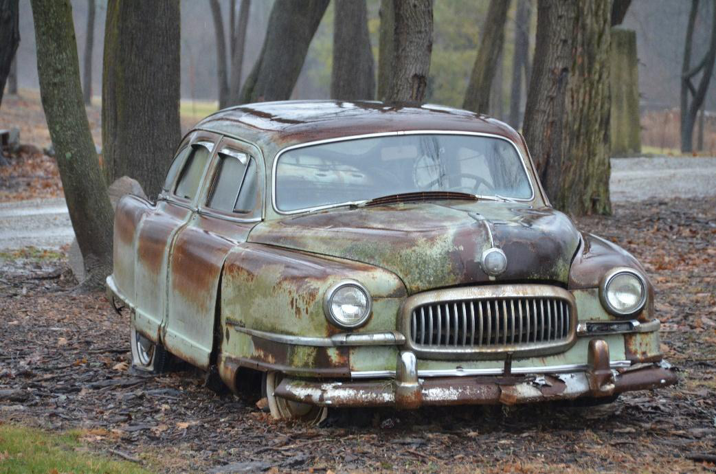 An old, rusty abandoned car with flat tires and a missing hubcap parked amongst tall trees in an overgrown area.