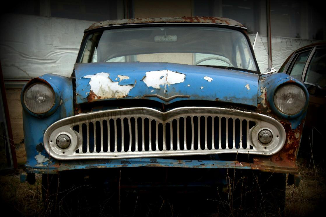 A blue vintage junk car with peeling paint and rust, featuring a prominent front grille and headlight.