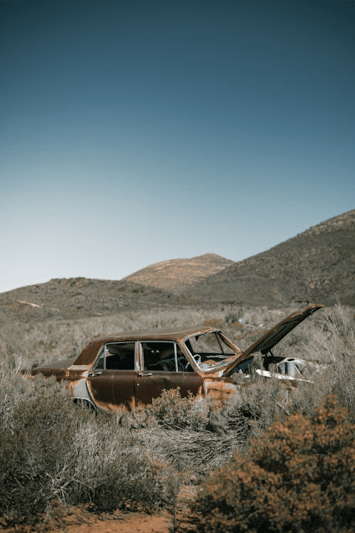 Rusted abandoned car with an open hood sitting in overgrown grass in a dry, mountainous landscape.