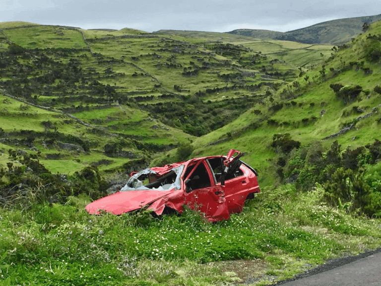 A severely damaged red car sits abandoned on a grassy hillside with rolling green hills in the background.