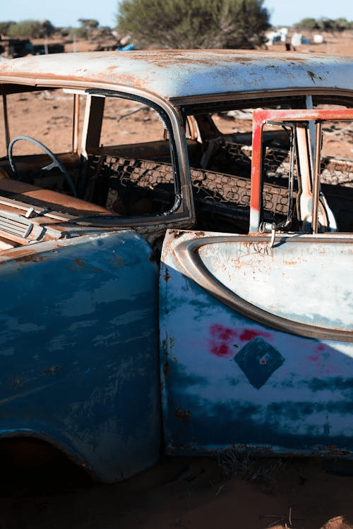 Close-up of a rusted, abandoned blue car with a partially open door in a dry, rural area.
