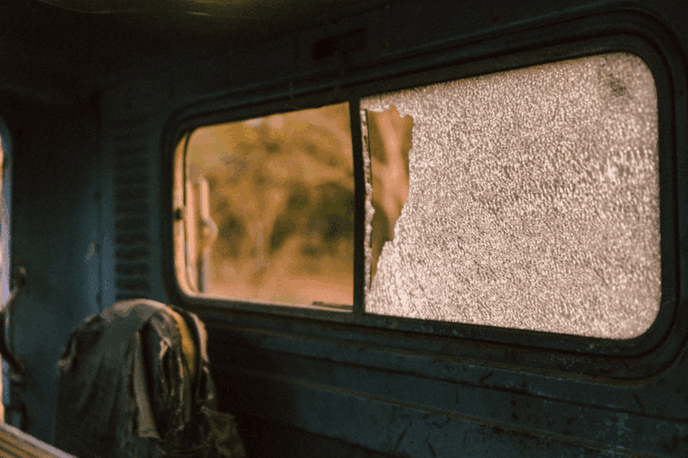Interior of an old vehicle with a shattered side window and worn seat, looking out onto a blurred outdoor background at sunset.