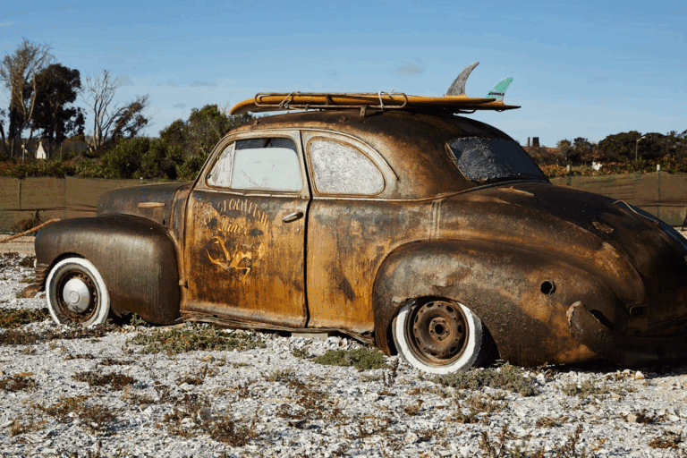 Rusted vintage car with flat tires and surfboards on the roof, abandoned on rocky ground under a clear sky.