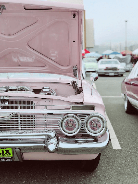 Close-up of a pink vintage car with its hood open, showing the engine area at an outdoor car show.
