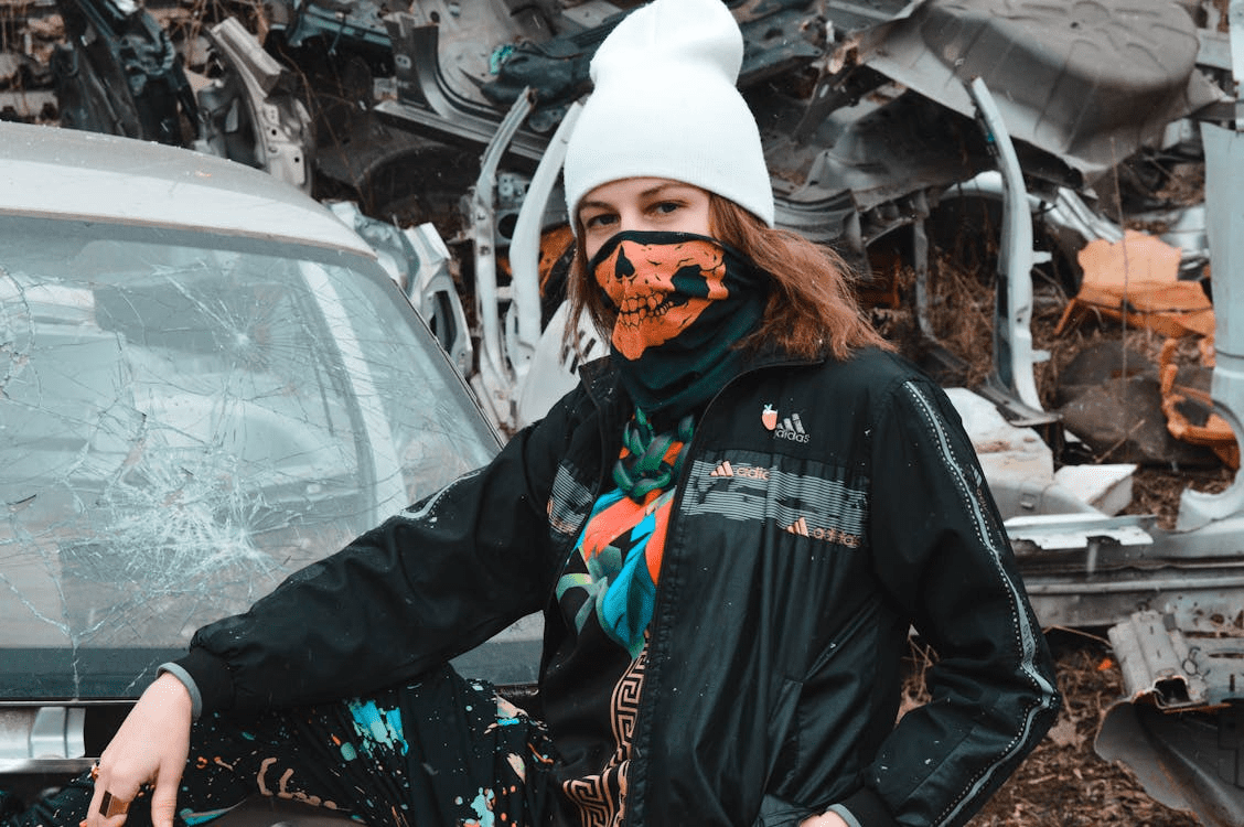 Person wearing a white beanie and orange skull face mask standing next to a damaged vehicle at a junkyard.